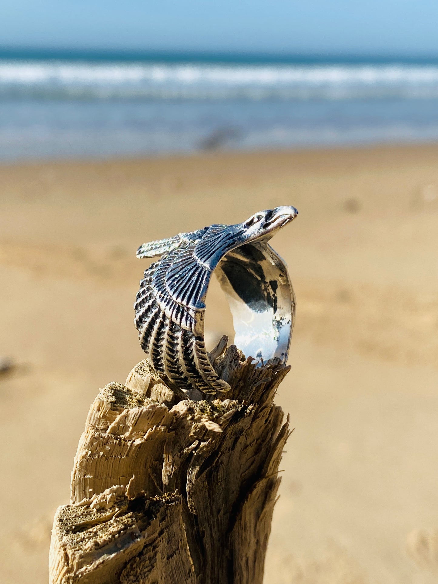bague en argent ajustable représentant un aigle posée sur un baton en bois sur la plage devant la mer