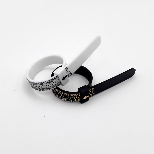 Photo de deux baguiers (Anneaux de mesure pour les tailles de bague) de couleur noire et blanche posés côte à côte sur un fond blanc. Les baguiers affichent des graduations de taille en chiffres dorés et noirs, avec une petite flèche indiquant la taille. Produit par la boutique de bijoux en argent "Altiplano".