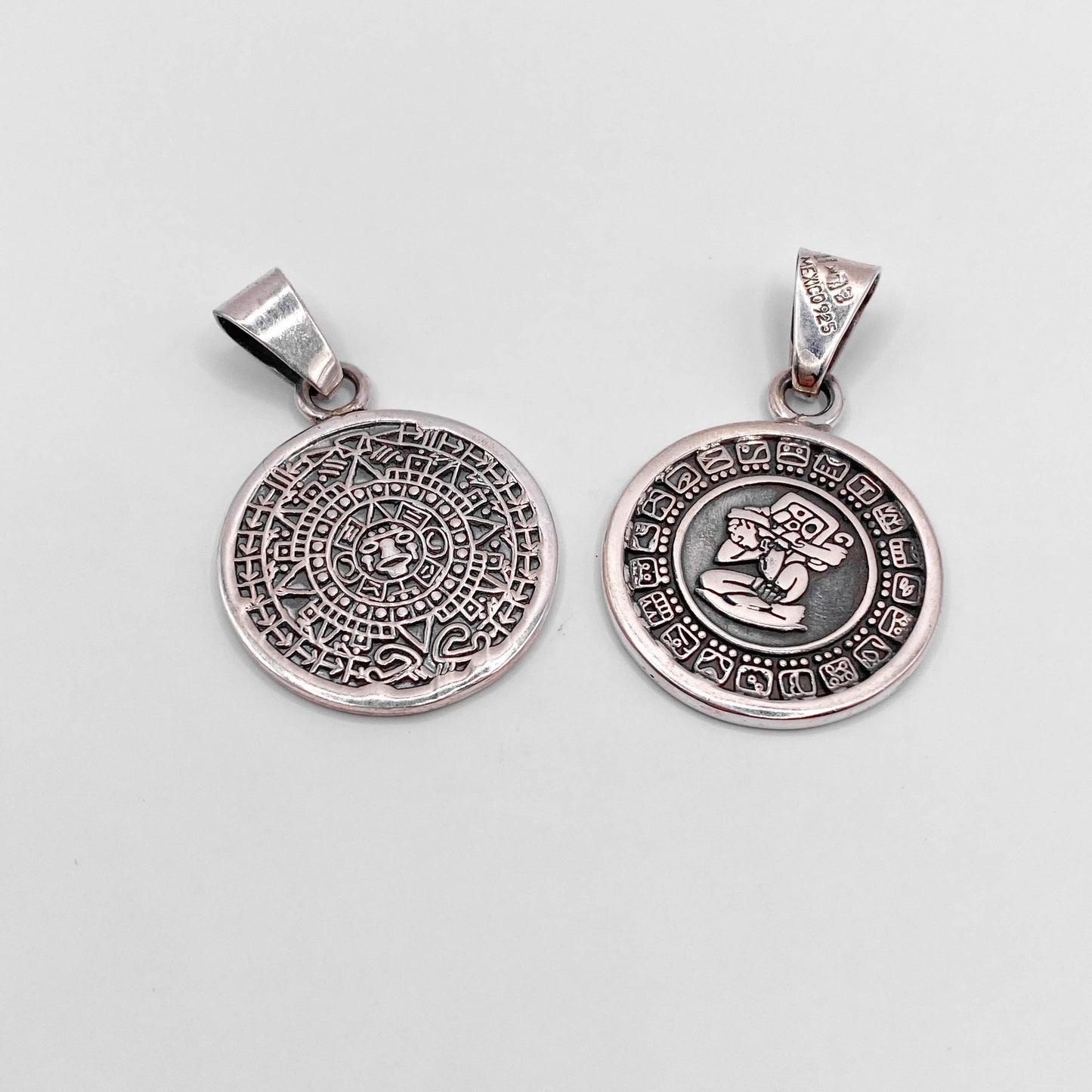 Deux pendentifs en argent à motifs aztèques disponibles sur la boutique en ligne Altiplano. Le premier pendentif présente un calendrier maya, tandis que le second met en avant un personnage gravé entouré de symboles mayas.