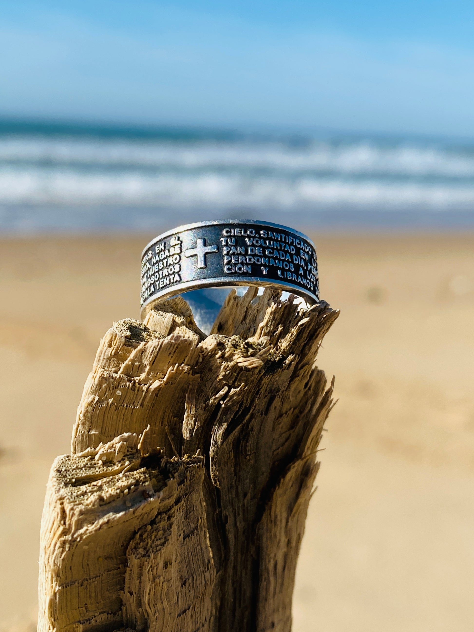 Bague en argent intitulée "PADRE" de la boutique Altiplano, ornée de gravures religieuses et d'une croix, posée sur un morceau de bois sur une plage avec l'océan en arrière-plan.