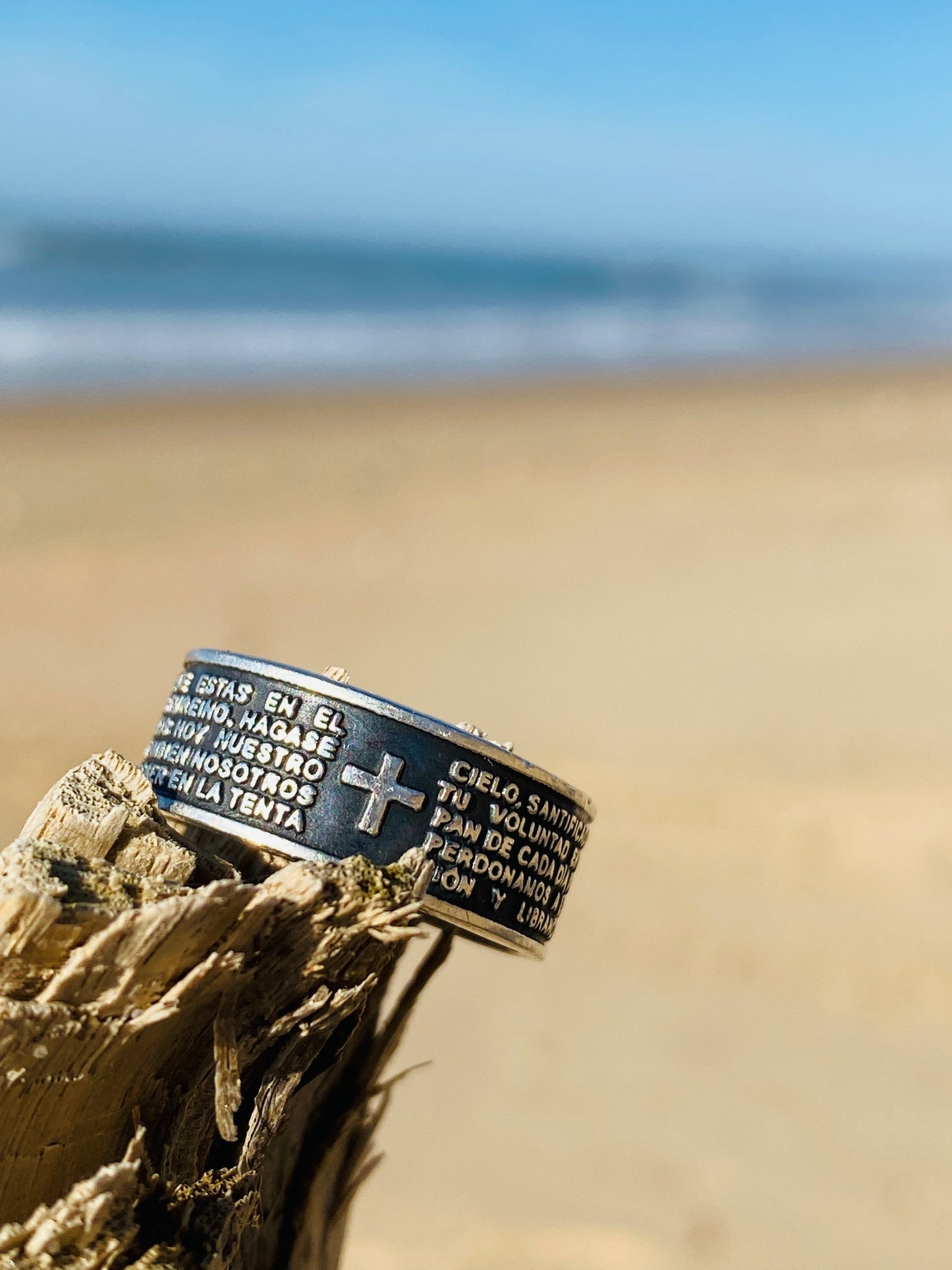 Bague en argent finement gravée intitulée "PADRE", provenant de la boutique Altiplano. Inspirée par l’artisanat latino-américain, cette pièce arbore une croix et un texte religieux, sur une plage, symbolisant foi et spiritualité.