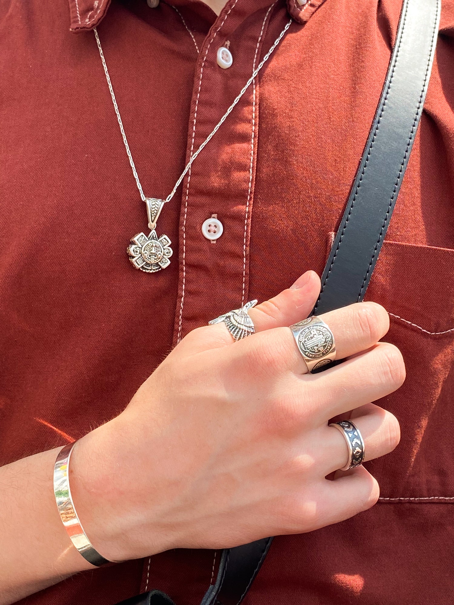 Personne portant divers bijoux en argent, comprenant un collier avec pendentif orné, plusieurs bagues et un bracelet simple. Les bijoux proviennent de la boutique en ligne Altiplano, spécialisée dans les bijoux en argent latino-américains. La chemise rouge brune met en valeur l'éclat du métal.