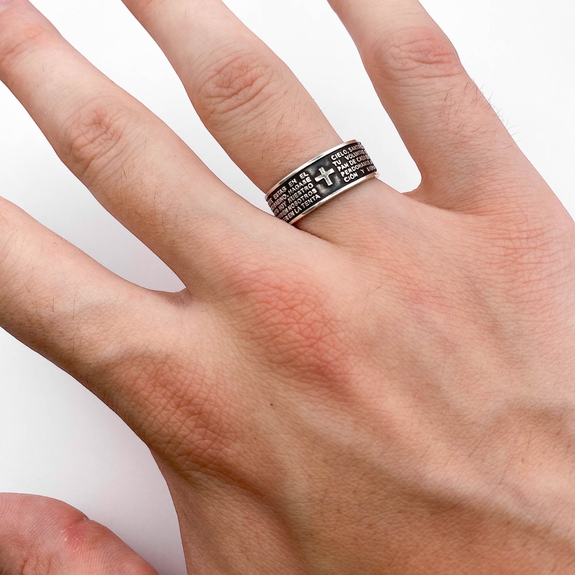 Bague en argent intitulée "PADRE" de la collection Altiplano, affichant des inscriptions et une croix sur le dessus. La bague est portée sur une main. Bijou élégant de style latino-américain.